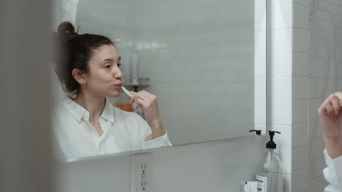 Adult woman brushing teeth, reflected in bathroom mirror. Clean and minimal style.